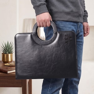 May include: A black rectangular briefcase with a smooth finish and a circular handle integrated into the top edge. The bag features a subtle embossed logo on the upper right corner. The design is structured with clean lines and a minimalist aesthetic.