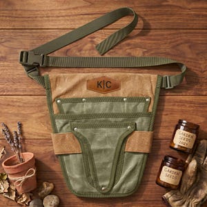 May include: A personalized garden tool belt in olive green and tan canvas with a brown leather patch. The belt has multiple pockets and an adjustable strap. Two jars of garden seeds and a pair of gardening gloves are also visible.