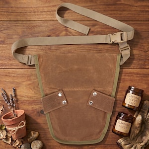 May include: A brown canvas gardening tool belt with a tan adjustable strap and buckle. The belt has two pockets with silver rivets and a green trim. Two jars labeled "Garden Seeds 2024" and a small terracotta pot with lavender are also visible.