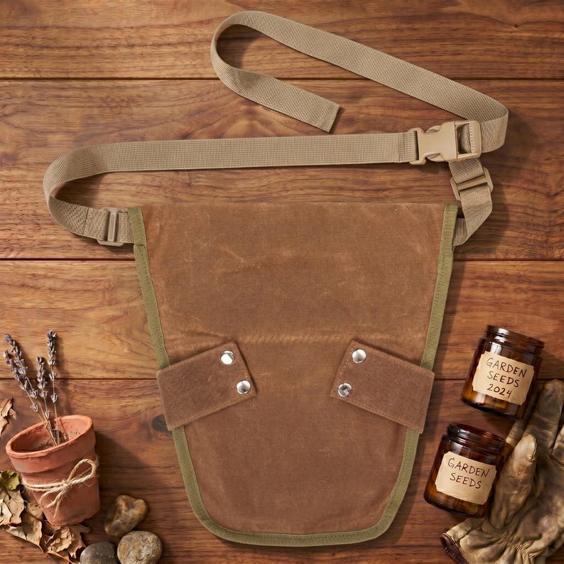 May include: A brown canvas gardening tool belt with a tan adjustable strap and buckle. The belt has two pockets with silver rivets and a green trim. Two jars labeled "Garden Seeds 2024" and a small terracotta pot with lavender are also visible.