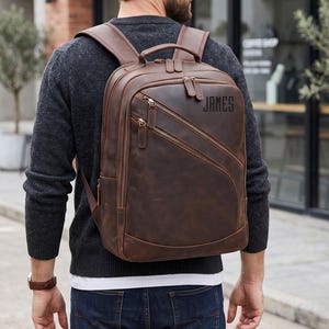 May include: A dark brown leather backpack featuring multiple zippered compartments, adjustable shoulder straps, and a top handle. The front of the bag is personalised with the name JAMES printed in a bold, dark, sans-serif font on the upper right side.