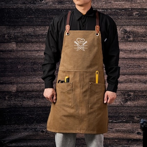 May include: Brown canvas apron with leather straps and metal hardware. The apron has two front pockets and a chest pocket. The apron has a chef's hat and crossed knives graphic with the name "James" printed below. The person is wearing a black long-sleeve shirt.