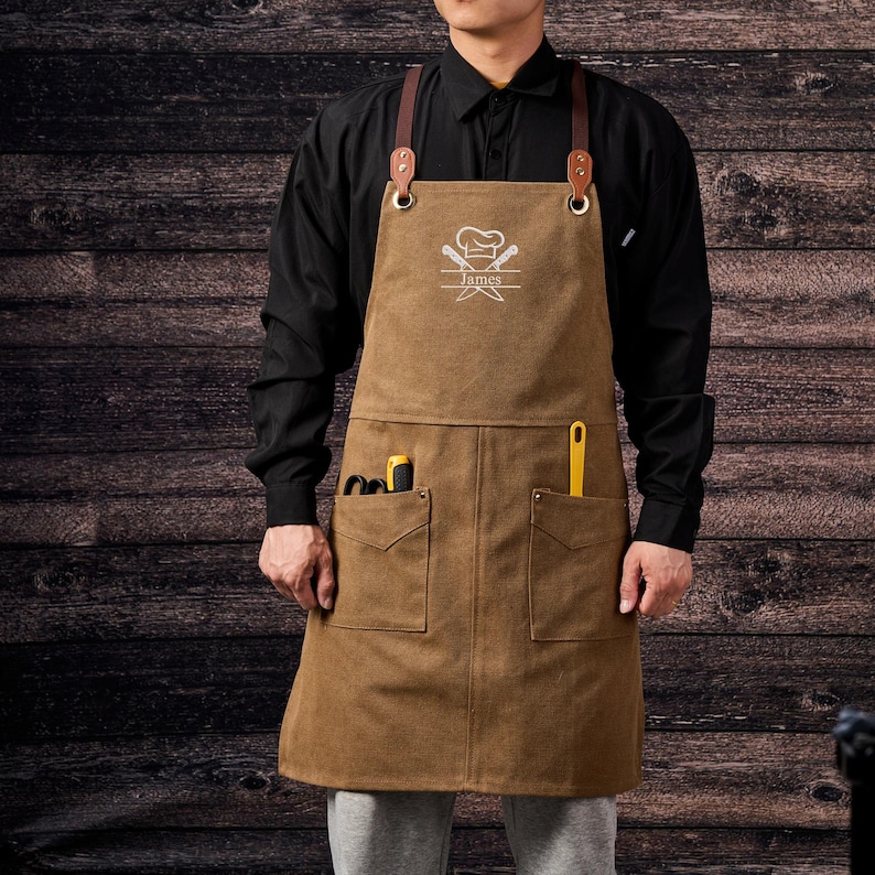 May include: Brown canvas apron with leather straps and metal hardware. The apron has two front pockets and a chest pocket. The apron has a chef's hat and crossed knives graphic with the name "James" printed below. The person is wearing a black long-sleeve shirt.