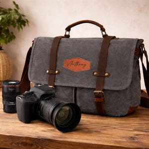 Könnte beinhalten: Graue Kameratasche aus Canvas mit braunen Lederakzenten, Riemen und Griff. Die Tasche hat eine Frontklappe und einen Lederaufnäher mit dem Namen "Anthony". Eine schwarze Kamera und ein Objektiv liegen auf einer Holzoberfläche.