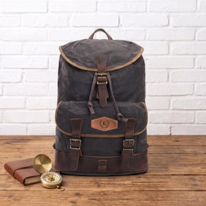 Könnte beinhalten: Ein Rucksack aus anthrazitfarbenem gewachstem Segeltuch mit braunen Lederbesätzen, einer oberen Klappe mit Schnallenverschluss und einer Vordertasche mit zwei Schnallenriemen. Ein Lederaufnäher auf der Vordertasche zeigt den Buchstaben K mit Lorbeerkranz.