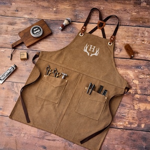 May include: A brown canvas apron with leather straps and pockets, featuring a white antler design and the letter "H". The apron is surrounded by barber tools, including scissors, a razor, and shaving cream, all arranged on a wooden surface.