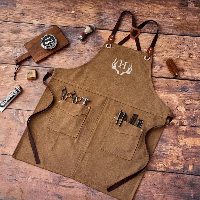 May include: A brown canvas apron with leather straps and pockets, featuring a white antler design and the letter "H". The apron is surrounded by barber tools, including scissors, a razor, and shaving cream, all arranged on a wooden surface.