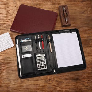 May include: A black leather portfolio organizer featuring a calculator, multiple pen loops, card slots, and a memo pad. A separate maroon leather folder and a wooden pen case with a silver pen are included in this professional stationery set.