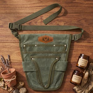 May include: A green canvas gardening tool belt with multiple pockets and an adjustable strap. A leather patch with a monogram is centered on the front. Two jars of garden seeds and a pair of gardening gloves are also visible.