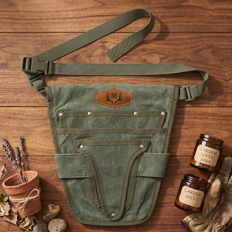May include: A green canvas gardening tool belt with multiple pockets and an adjustable strap. A leather patch with a monogram is centered on the front. Two jars of garden seeds and a pair of gardening gloves are also visible.