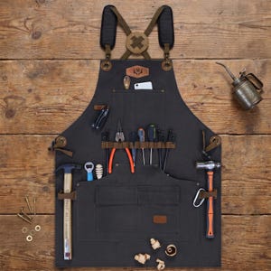 May include: A dark gray canvas work apron featuring brown leather straps, a cross-back design, and multiple pockets. The apron is equipped with various tools including a hammer, pliers, screwdrivers, and a metal oil can, all organized in loops and pockets.