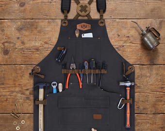 Puede incluir: Un delantal de trabajo de lona gris oscuro con correas de cuero marrón, diseño de espalda cruzada y múltiples bolsillos. El delantal está equipado con varias herramientas, incluyendo un martillo, alicates, destornilladores y una aceitera de metal.