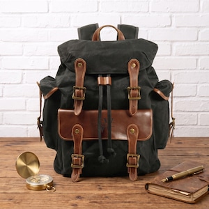 May include: A dark charcoal gray backpack featuring brown leather straps, brass-toned buckles, and a front pocket flap. The bag has a drawstring closure, side pockets, and a top handle. A gold compass and a brown leather journal with a pen are nearby.