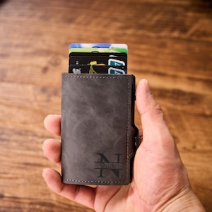 Puede incluir: Una cartera de cuero gris con un monograma de las letras "N" e "I" y el nombre "Nick" debajo. La cartera está llena de varias tarjetas de crédito y se sostiene en una mano.