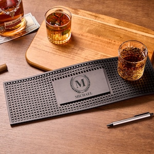 Può includere: Una scena da bar con una bottiglia di whisky, due bicchieri di liquido ambrato e un sigaro. Al centro c'è un tappetino da bar nero con una targhetta personalizzata "MICHAEL". Sono presenti anche un tagliere in legno e una penna.