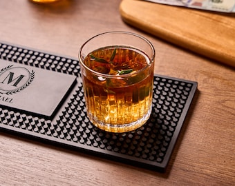 May include: A rocks glass filled with a golden-brown beverage and ice sits on a black bar mat. The mat has a repeating pattern and a personalized nameplate with the name "MICHAEL" and a monogram. A wooden cutting board is in the background.