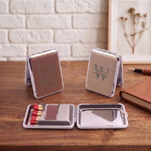 Puede incluir: Dos cajas de cerillas rectangulares de metal con tapa abatible, una marrón y otra beige, con monogramas grabados. La caja abierta muestra una fila de cerillas de madera con puntas rojas junto a una superficie de rascado oscura en el interior.
