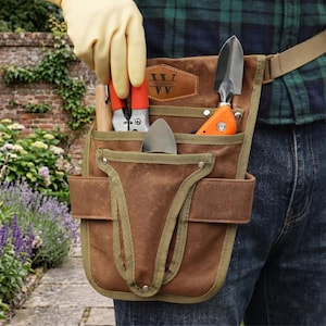May include: A brown canvas gardening tool belt with green trim, holding a trowel, pruning shears, and a small shovel. The belt has a personalized leather patch with the name "William". The person is wearing a plaid shirt and jeans.