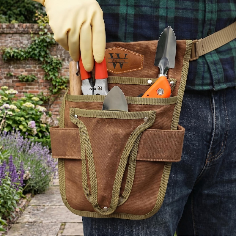 May include: A brown canvas gardening tool belt with green trim, holding a trowel, pruning shears, and a small shovel. The belt has a personalized leather patch with the name "William". The person is wearing a plaid shirt and jeans.