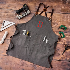 May include: A gray canvas apron with brown leather straps and pockets, embroidered with a red and blue letter "D". Tools including a staple gun, wrench, and clamps are arranged around the apron on a wooden surface.