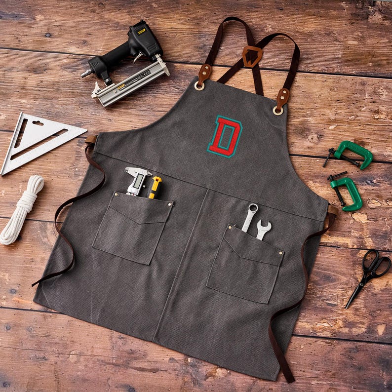 May include: A gray canvas apron with brown leather straps and pockets, embroidered with a red and blue letter "D". Tools including a staple gun, wrench, and clamps are arranged around the apron on a wooden surface.