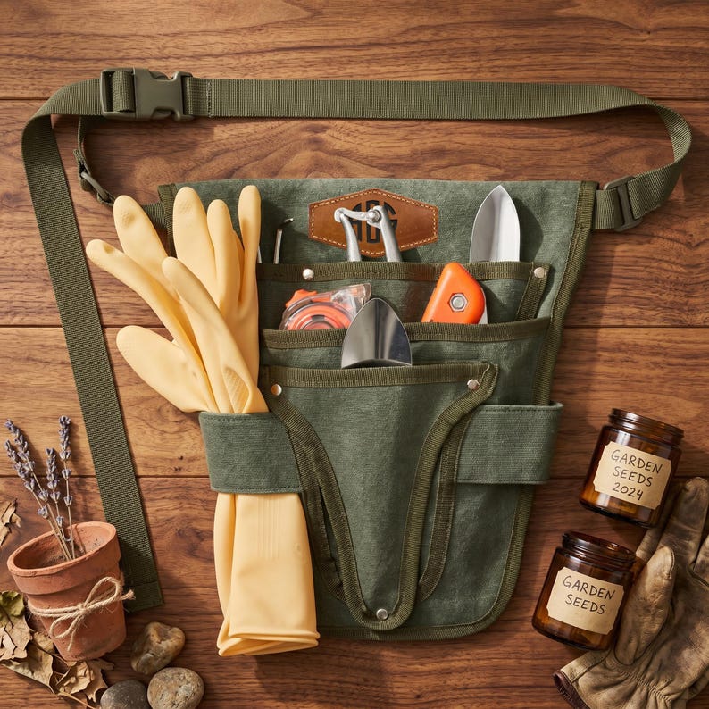 May include: A green canvas gardening tool belt with multiple pockets, filled with tools, gloves, and a tape measure. Includes two jars of garden seeds and a small terracotta pot with lavender. The belt has an adjustable strap.