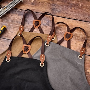 May include: Close-up of four aprons in black, tan, and gray. Each apron has brown leather straps and accents, with adjustable cross-back design. The aprons are made of durable fabric and feature brass-colored metal hardware.