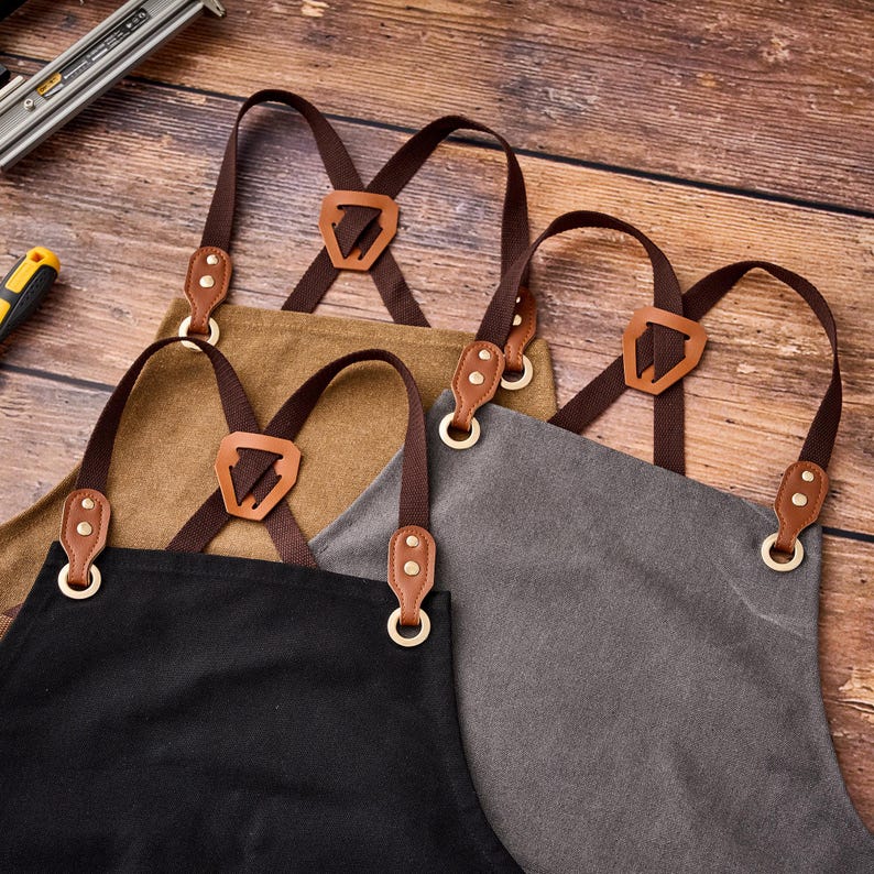 May include: Close-up of four aprons in black, tan, and gray. Each apron has brown leather straps and accents, with adjustable cross-back design. The aprons are made of durable fabric and feature brass-colored metal hardware.
