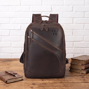 Könnte beinhalten: Ein dunkelbrauner Lederrucksack mit zwei diagonalen Reißverschlusstaschen auf der Vorderseite, einem Tragegriff und verstellbaren Schultergurten. Auf der oberen Vorderseite ist der Buchstabe W und der Name William zwischen zwei Linien eingraviert.