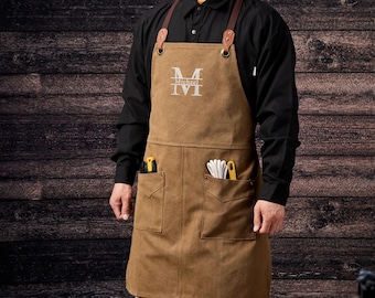May include: Brown canvas apron with leather straps and pockets. The apron has the letter "M" and the name "Michael" embroidered on the chest. Tools are visible in the pockets. The apron is worn over a black shirt.