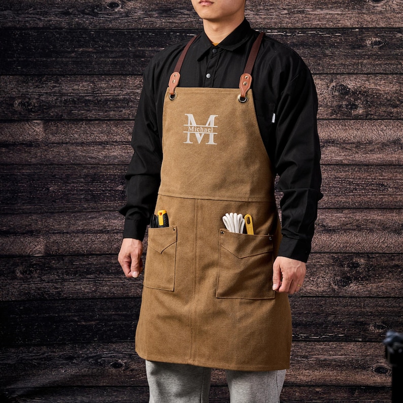 May include: Brown canvas apron with leather straps and pockets. The apron has the letter "M" and the name "Michael" embroidered on the chest. Tools are visible in the pockets. The apron is worn over a black shirt.