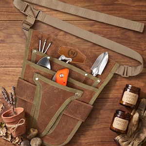 May include: A brown canvas gardening tool belt with multiple pockets, holding various metal tools and an orange handled knife. The belt has a tan strap and buckle. Two small jars of garden seeds and a small terracotta pot with lavender are also visible.