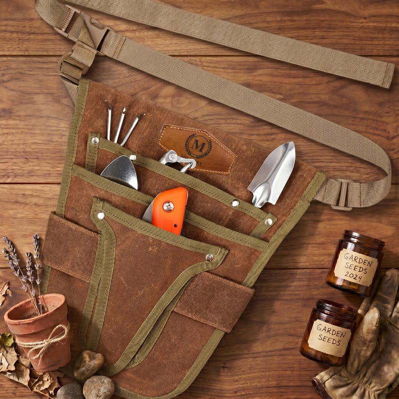 May include: A brown canvas gardening tool belt with multiple pockets, holding various metal tools and an orange handled knife. The belt has a tan strap and buckle. Two small jars of garden seeds and a small terracotta pot with lavender are also visible.