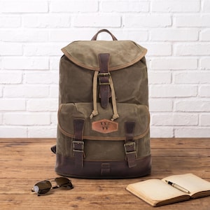 Könnte beinhalten: Ein olivgrüner Rucksack aus Segeltuch mit einem Boden, einem oberen Klappdeckel und Riemen aus dunkelbraunem Leder. Die Tasche verfügt über eine Vordertasche mit einem Lederaufnäher mit dem Buchstaben W, Messingschnallen und einen Kordelzug.