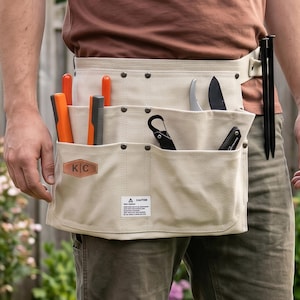 Peut inclure: Une ceinture porte-outils en toile beige dotée de multiples poches et d'une sangle. Elle comporte un écusson en cuir marron avec les lettres K|C, une étiquette d'avertissement blanche et divers outils comme des manches orange et des lames noires.