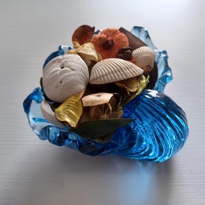 May include: A decorative blue glass bowl filled with seashells, dried flowers, and potpourri. The bowl has a wavy, shell-like design. The contents include white and tan seashells, orange flowers, and dried leaves.