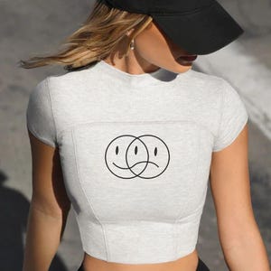 May include: A light gray crop top with a black graphic of two overlapping smiley faces, one happy and one sad. The model is wearing a black baseball cap with white text. The top has a fitted design.
