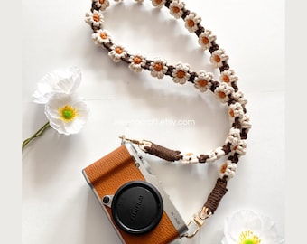 May include: A camera strap featuring a series of crocheted daisy flowers in cream and orange, connected by a dark brown cord. The strap is attached to a tan and silver Fujifilm X-A2 camera with a black lens cap. The text Jesenocraft.etsy.com is visible.