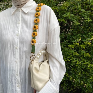 May include: A cream-colored handbag with a decorative strap featuring crocheted yellow sunflowers with brown centers and green leaves. The strap is attached to the bag with gold-colored hardware. The text "Jesenacraft.etsy.com" is visible on the strap.