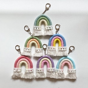 May include: A set of seven rainbow-coloured crocheted keychains with white fringe. Each keychain features a rainbow arch with a white lace trim and a silver key ring.