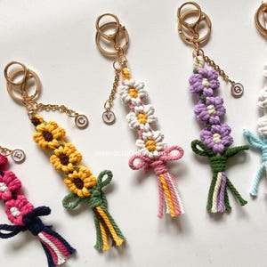 May include: Macrame keychains with flower designs in yellow, white, and purple. Each keychain has a gold-colored clasp and chain, with a tassel at the bottom. The keychains are handmade and feature a variety of floral patterns.