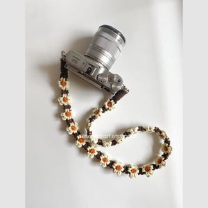 May include: A silver digital camera with a brown and cream daisy chain camera strap. The strap is made of crocheted flowers with orange centers. The camera is on a white surface.