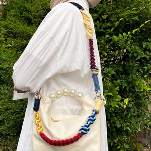 May include: Cream-colored handbag with a pearl handle and a colorful, braided strap. The strap features sections of navy, yellow, red, and blue. The bag has a zipper closure and the text "Jesenacraft.etsy.com" is visible.