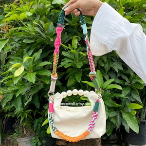 May include: Cream-coloured handbag with a pearl handle and a colourful macrame strap. The strap features pink, green, orange, and blue accents. The bag is held against a backdrop of green foliage.