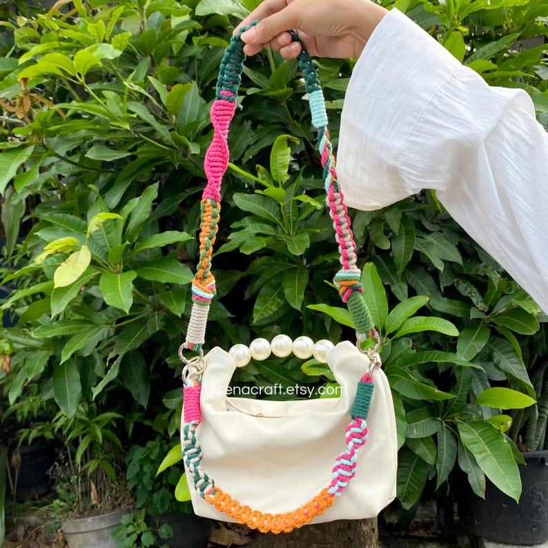 May include: Cream-coloured handbag with a pearl handle and a colourful macrame strap. The strap features pink, green, orange, and blue accents. The bag is held against a backdrop of green foliage.