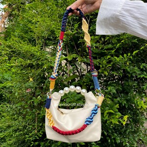 May include: Cream-colored handbag with a colorful braided strap. The strap features blue, red, yellow, and white accents. The bag has a pearl handle and a zipper closure. The Etsy shop name, desenacraft.etsy.com, is visible on the bag.