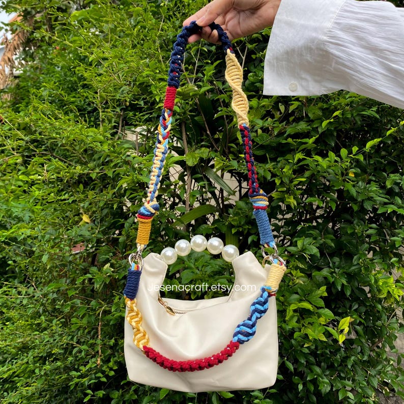 May include: Cream-colored handbag with a colorful braided strap. The strap features blue, red, yellow, and white accents. The bag has a pearl handle and a zipper closure. The Etsy shop name, desenacraft.etsy.com, is visible on the bag.