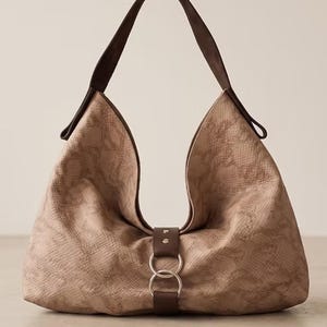 Pode incluir: Uma bolsa de ombro estilo hobo em bege claro com um padrão texturizado semelhante a pele de cobra. A bolsa apresenta uma alça castanho-escura e um fecho central de aba castanho-escuro fixado com dois anéis de metal prateados.