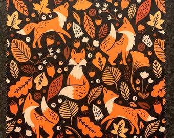 Puede incluir: Una tela negra con un patrón repetido de zorros naranjas y hojas de otoño. Los zorros son estilizados y tienen acentos blancos. Las hojas son en tonos de naranja, marrón y amarillo.
