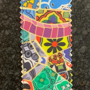 Puede incluir: Un marcapáginas colorido con un patrón de mosaico que presenta formas geométricas, flores y remolinos en tonos de azul, verde, amarillo, naranja, morado y rojo.